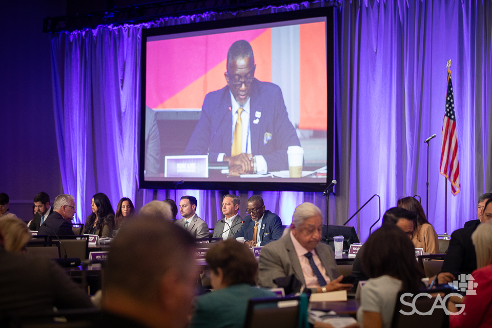 Conference with attendees seated, large screen displays a speaker, U.S. flag in background.
