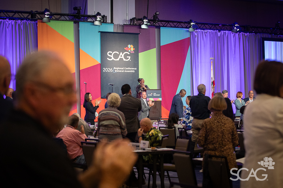 Conference audience facing colorful stage with speaker, some standing.