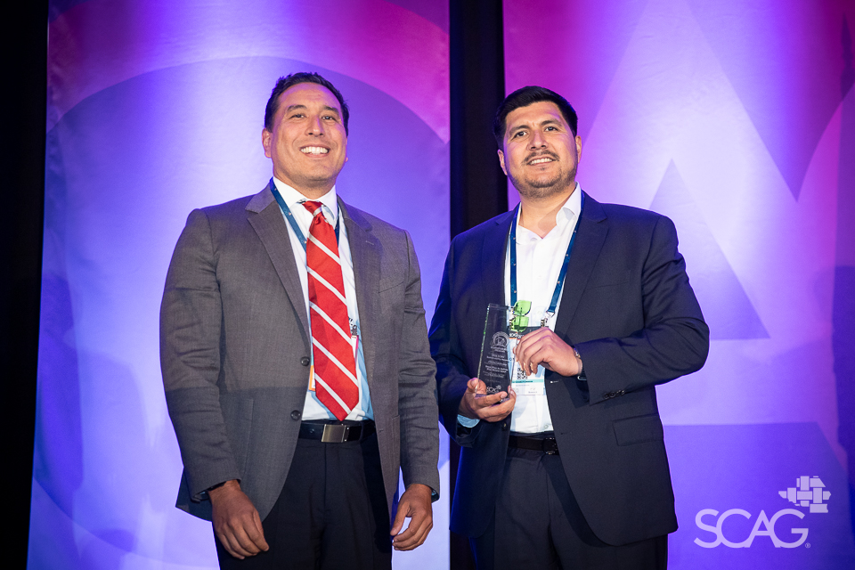 Two men in suits, one holding an award, stand against a blue and purple backdrop.