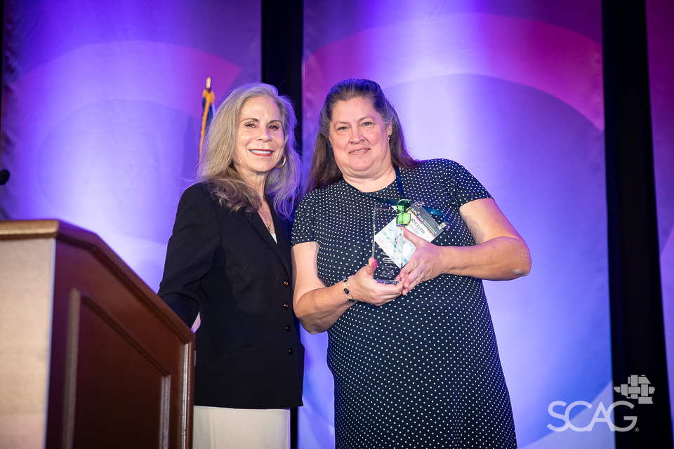 Two women stand together, one holding a glass award, against a purple backdrop.