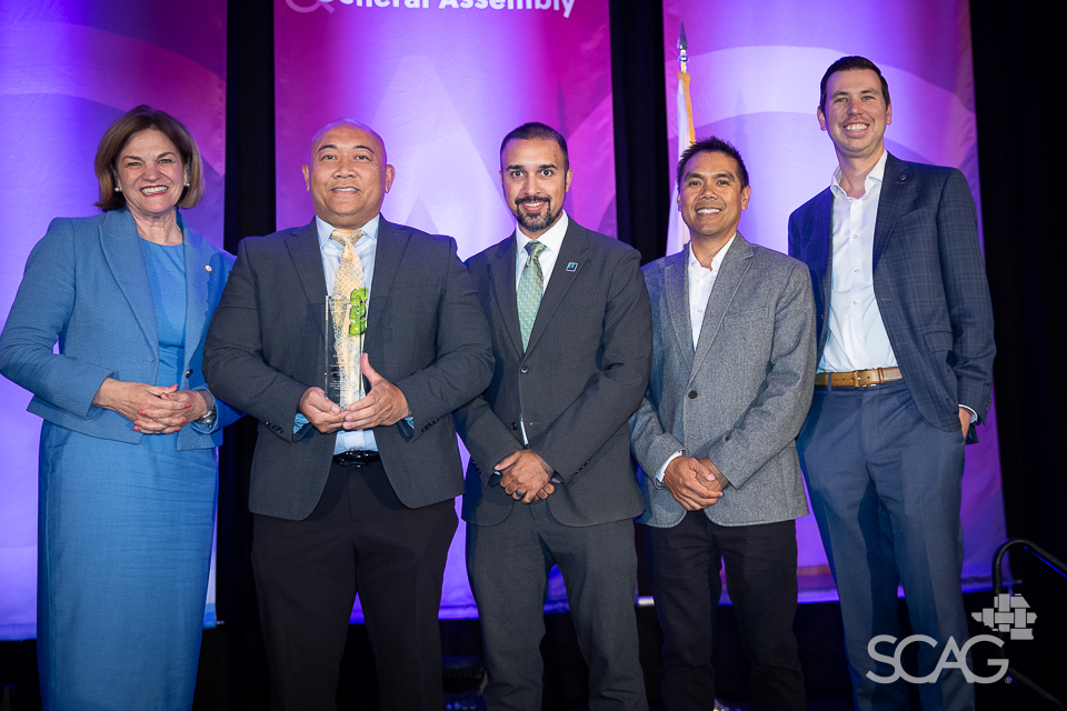 Five people in formal attire smiling on a stage with a purple backdrop.