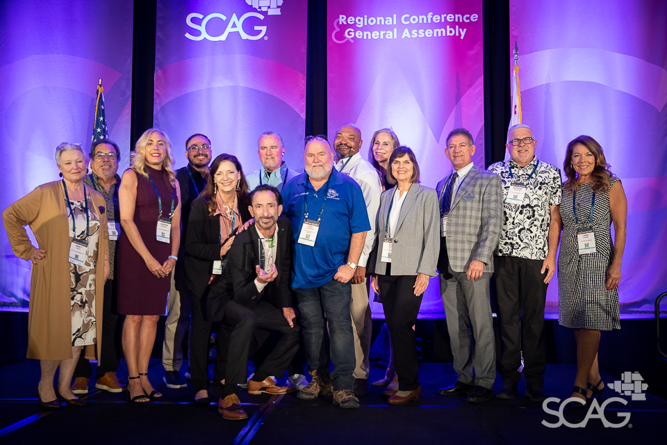 Group of people smiling on stage at a conference with purple backdrop.