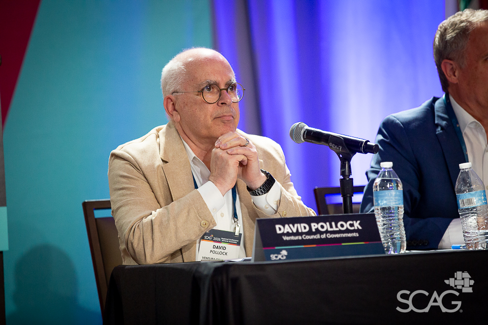 Seated man in a beige suit at a conference, appears thoughtful.
