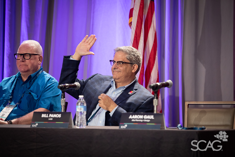 Two men seated at a panel, one raising his hand, with a U.S. flag behind them.