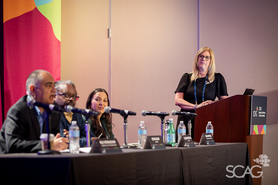 Panel discussion with four seated speakers and a woman standing at a podium.