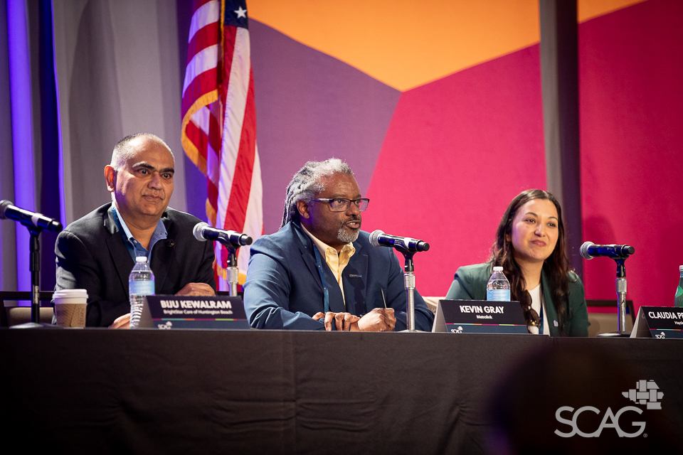 Panel of three speakers seated with microphones in front of a colorful backdrop and a flag.