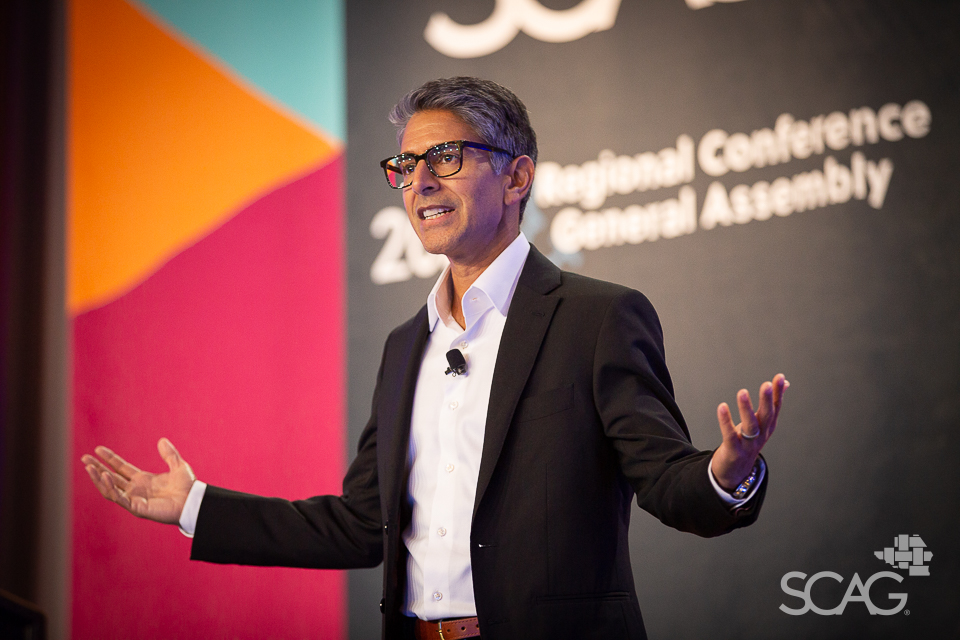 Man speaking energetically on stage at a conference, colorful backdrop.