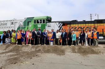 People in formal and safety attire stand with shovels on a dirt mound in front of two colorful trains at a groundbreaking ceremony under an overcast sky.