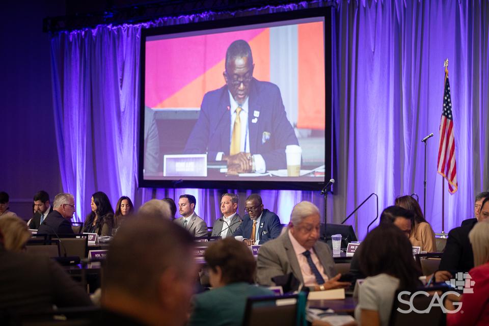 Conference with attendees seated, large screen displays a speaker, U.S. flag in background.