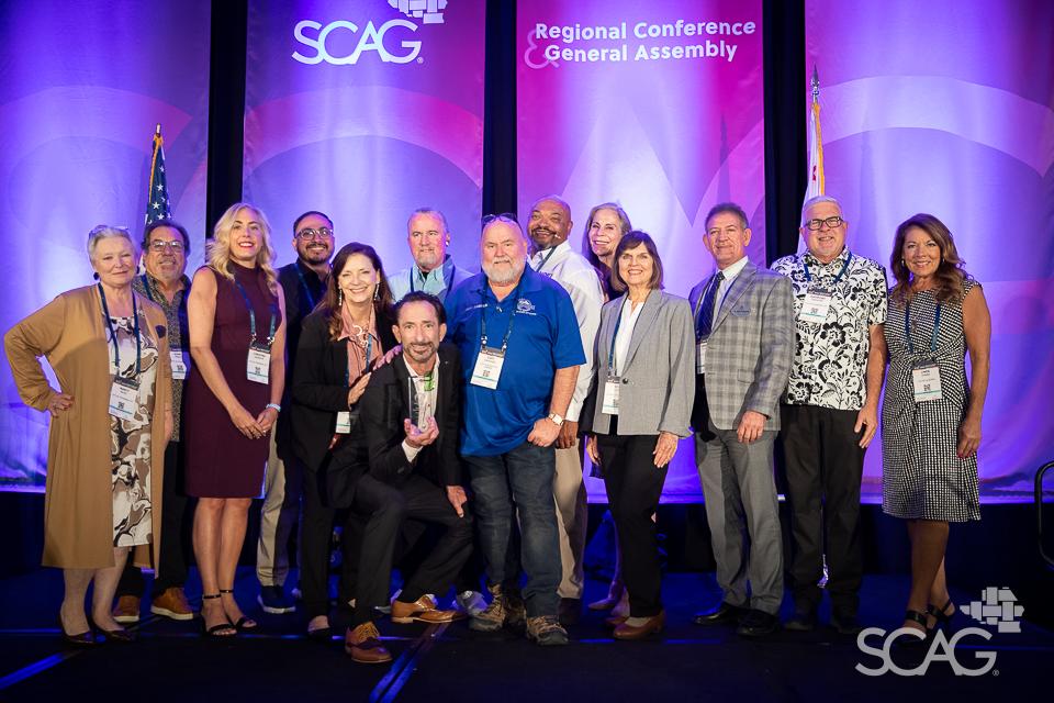 Group of people smiling on stage at a conference with purple backdrop.