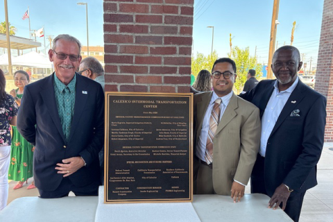 Three men smiling beside a commemorative plaque mounted on a brick pillar.