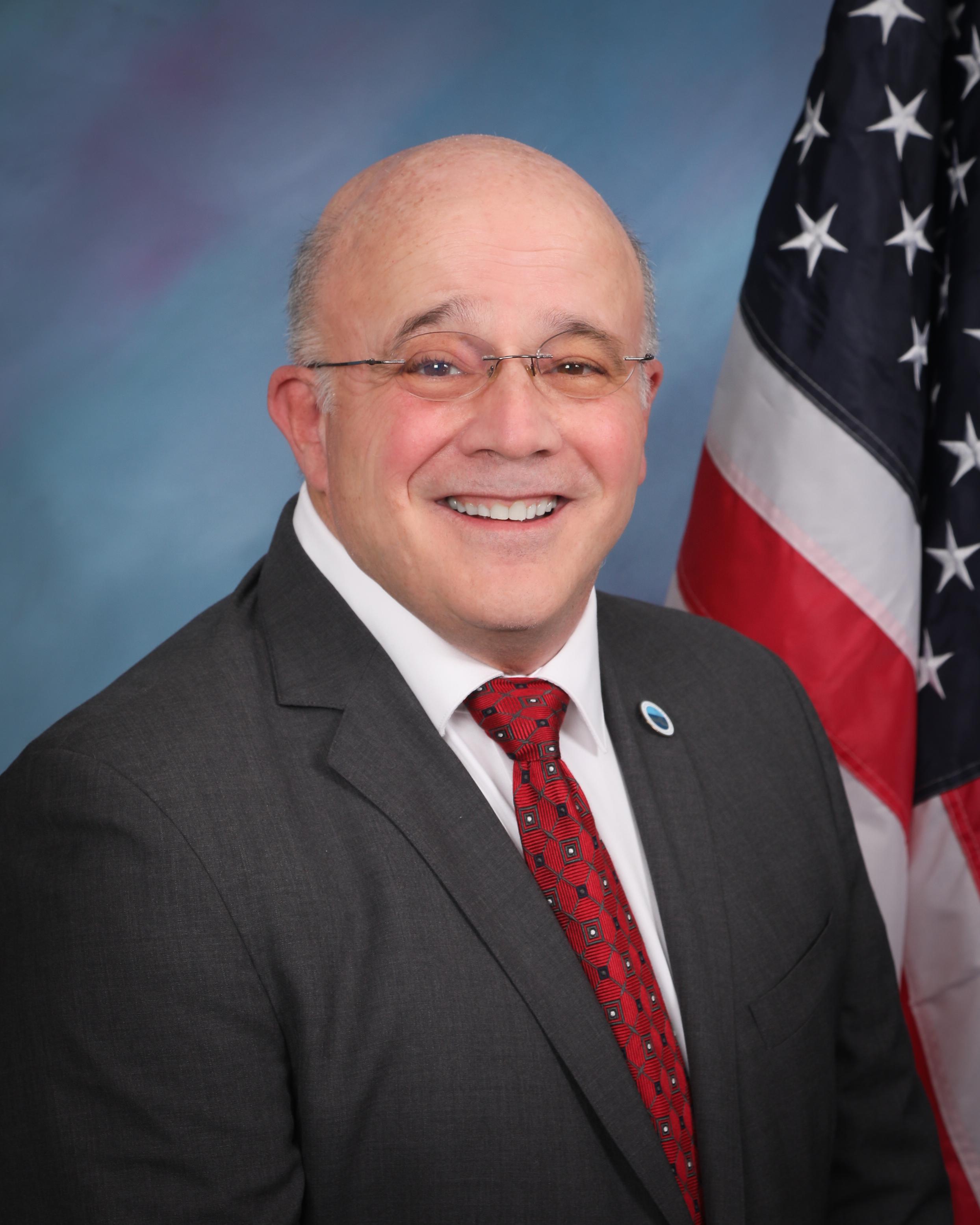 Bald man in a suit and red tie, smiling, with a U.S. flag backdrop.