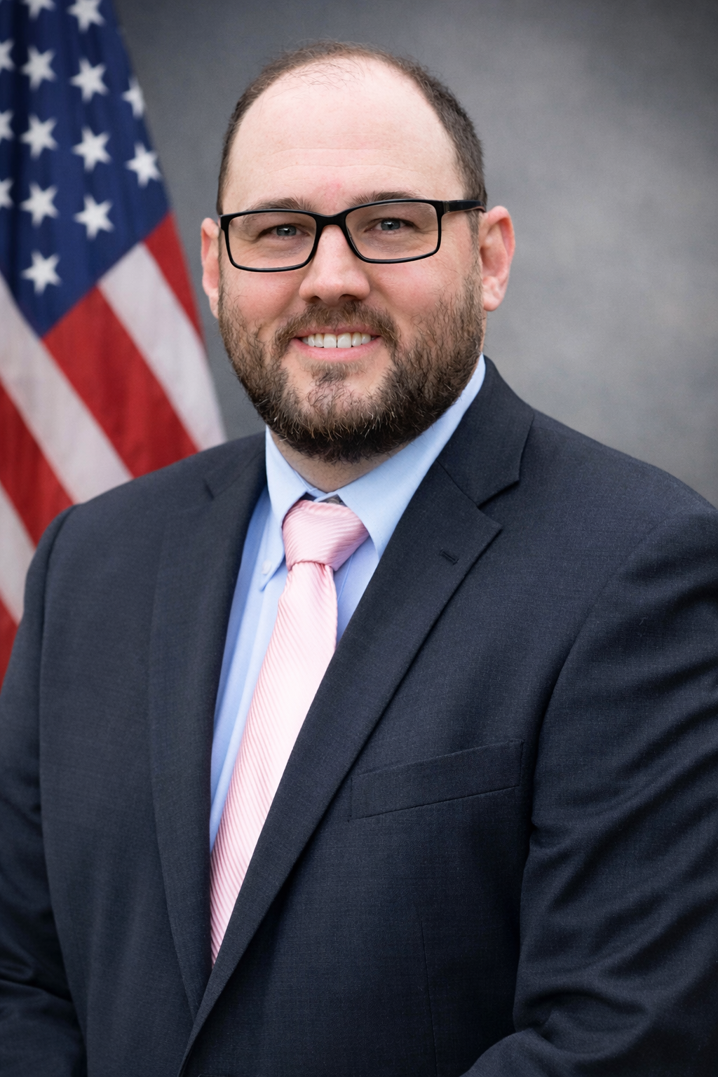 Man in suit with pink tie, glasses, and beard, standing in front of U.S. flag.