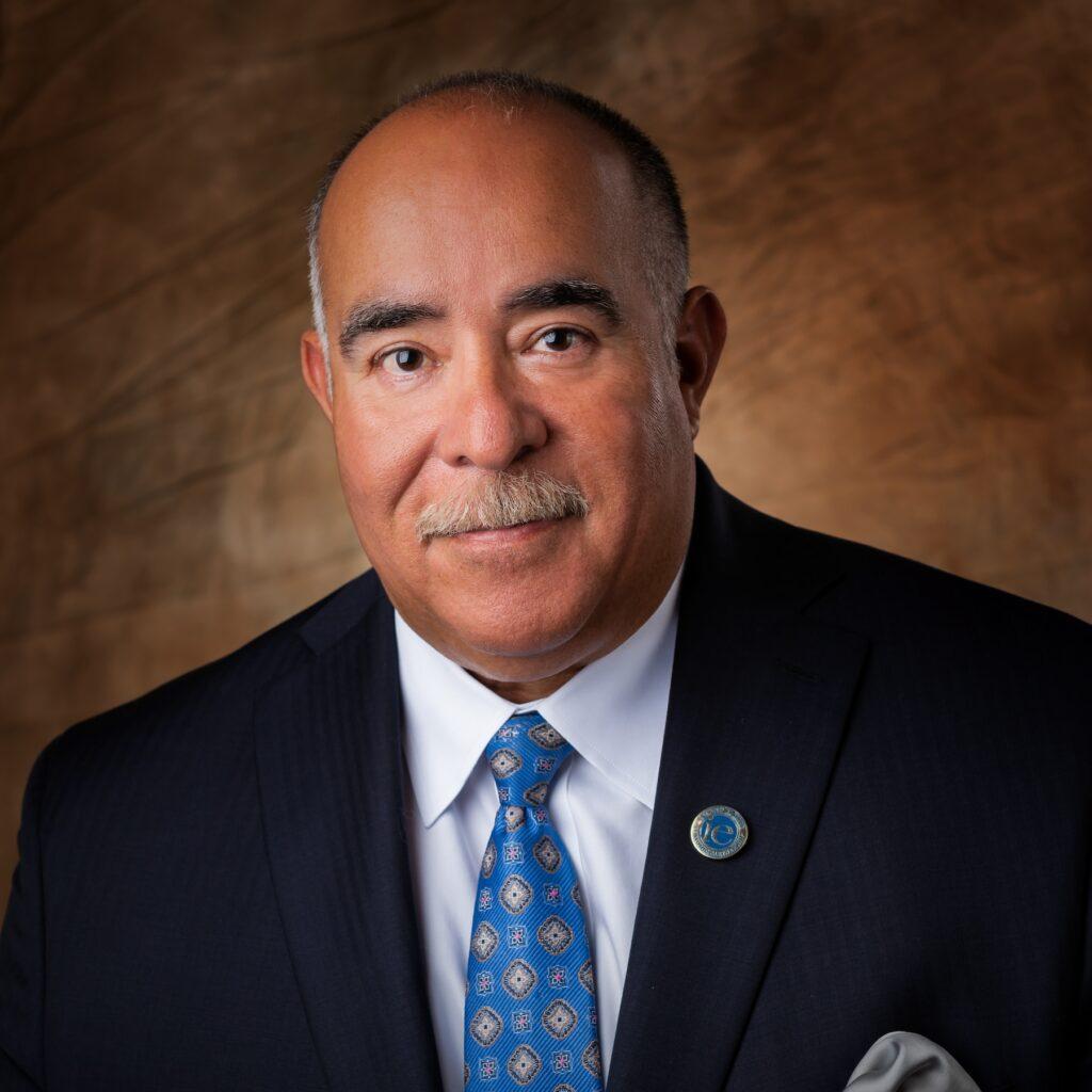 Man in a suit and tie, with a brown background, looking directly at the camera.