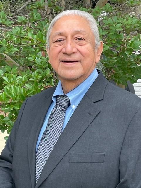 Elderly man in a suit smiles outdoors with greenery in the background.