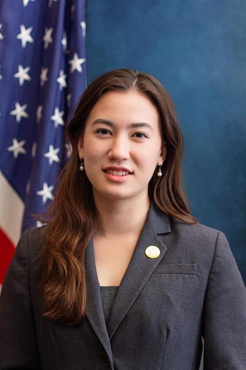 Woman in a suit with long hair stands beside a U.S. flag.